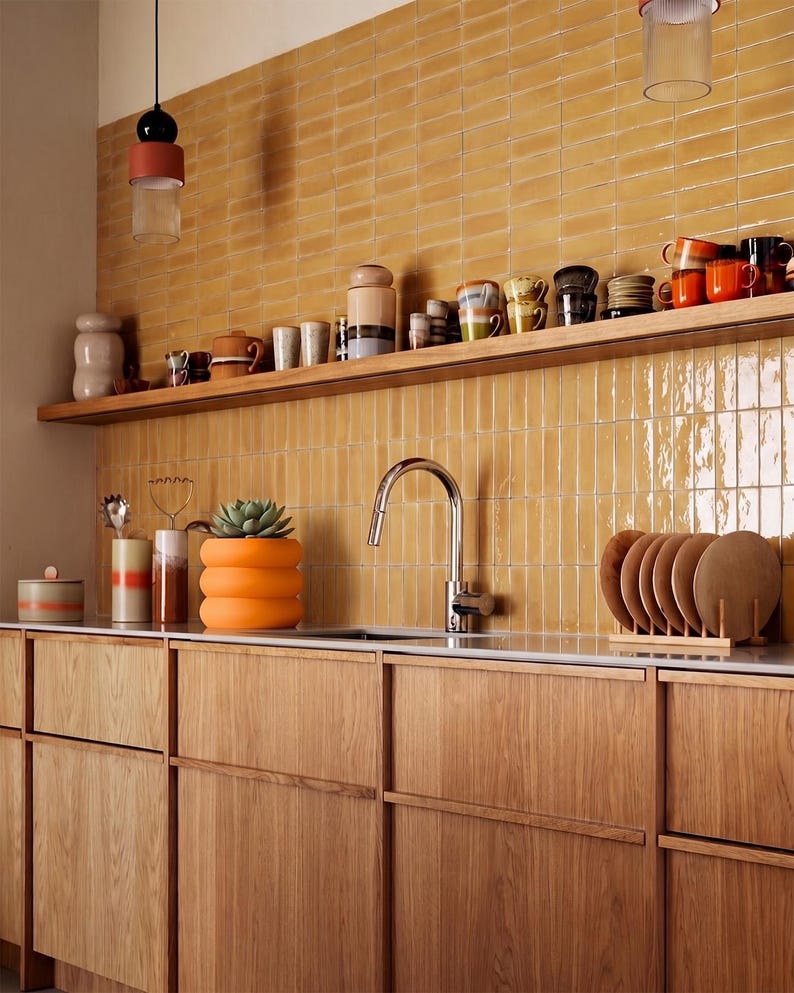 May include: A kitchen featuring light wood cabinets, a white countertop, and a backsplash of glossy mustard yellow rectangular tiles. A wooden shelf holds various ceramic mugs and containers, while a silver faucet sits above the sink.