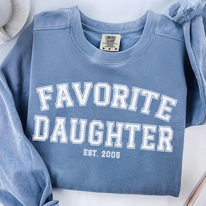 Puede incluir: Una sudadera azul vaquera con las palabras "FAVORITE DAUGHTER" en letras blancas. El texto "EST. 2005" est&aacute; debajo. La sudadera est&aacute; doblada, con una taza de caf&eacute; y un libro en el fondo.