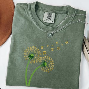 May include: A moss green t-shirt featuring a dandelion design with yellow pi symbols floating away. The shirt has a crew neck and short sleeves. A silver necklace is draped across the shirt. The word "MOSS" is printed at the bottom left.