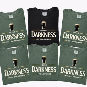 May include: A collection of six t-shirts, in black and moss green, with the phrase "Hello Darkness My Old Friend" printed in white and gold. Each shirt features a pint glass graphic above the text.