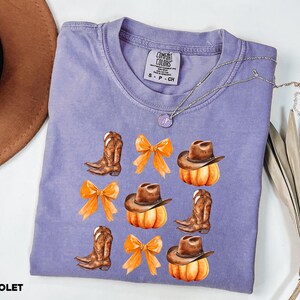 May include: A violet short-sleeve t-shirt with a Comfort Colors label featuring a graphic print of brown cowboy boots, orange bows, and pumpkins wearing brown cowboy hats. The shirt is made of 100% cotton and is size small.