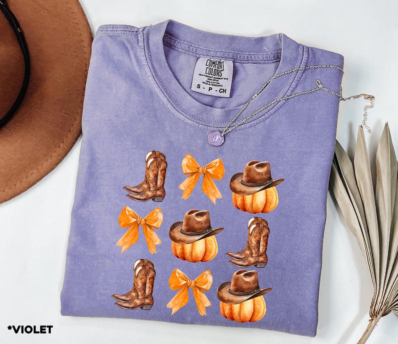 May include: A violet short-sleeve t-shirt with a Comfort Colors label featuring a graphic print of brown cowboy boots, orange bows, and pumpkins wearing brown cowboy hats. The shirt is made of 100% cotton and is size small.