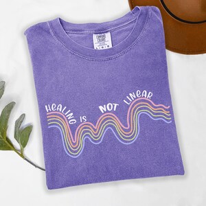 May include: Purple t-shirt with a white graphic that says "Healing is not linear" with a rainbow wave design.