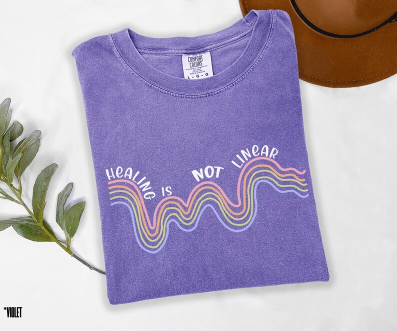 May include: Purple t-shirt with a white graphic that says "Healing is not linear" with a rainbow wave design.