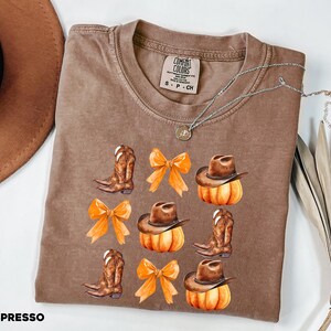 May include: A brown crewneck t-shirt featuring a graphic print of brown cowboy boots, orange bows, and brown cowboy hats resting on orange pumpkins. The inner neck label reads Comfort Colors, size S, 100% cotton, made in Honduras.