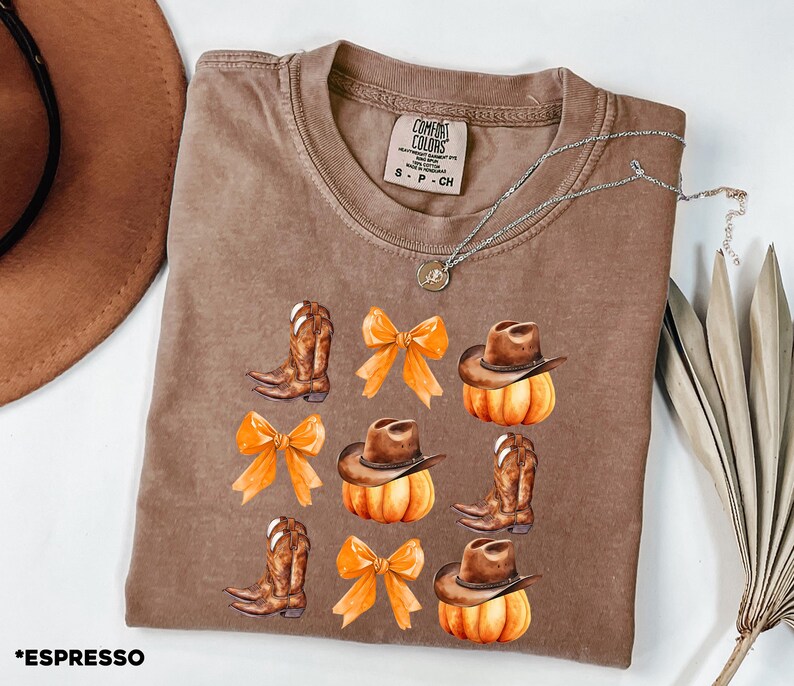 May include: A brown crewneck t-shirt featuring a graphic print of brown cowboy boots, orange bows, and brown cowboy hats resting on orange pumpkins. The inner neck label reads Comfort Colors, size S, 100% cotton, made in Honduras.