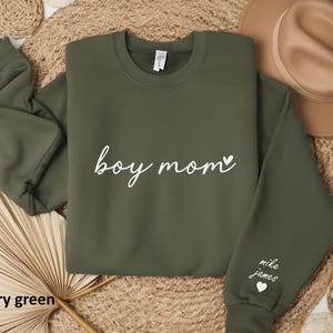 Op de afbeelding: Een olijfgroen sweatshirt met de tekst "boy mom" in witte cursieve letters en een klein hartje. De rechtermouw heeft de tekst "mike james" in witte letters en een klein hartje.