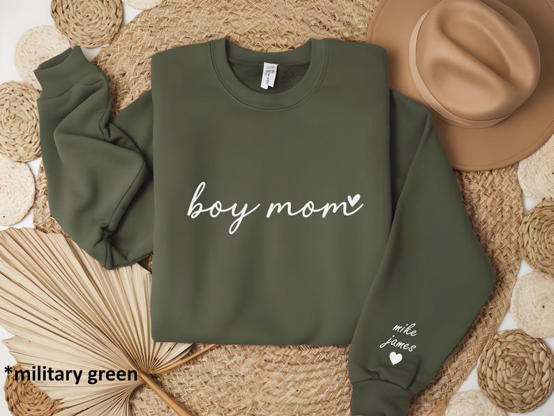 Op de afbeelding: Een olijfgroen sweatshirt met de tekst "boy mom" in witte cursieve letters en een klein hartje. De rechtermouw heeft de tekst "mike james" in witte letters en een klein hartje.