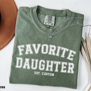 Puede incluir: Una camiseta verde musgo con las palabras "FAVORITE DAUGHTER" en letras blancas. La camiseta tambi&eacute;n tiene "EST. CUSTOM" impreso debajo del texto principal. Un collar plateado con un peque&ntilde;o colgante descansa sobre la camiseta.