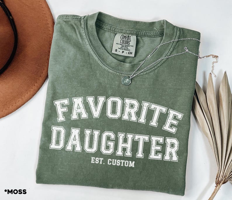 Puede incluir: Una camiseta verde musgo con las palabras "FAVORITE DAUGHTER" en letras blancas. La camiseta tambi&eacute;n tiene "EST. CUSTOM" impreso debajo del texto principal. Un collar plateado con un peque&ntilde;o colgante descansa sobre la camiseta.
