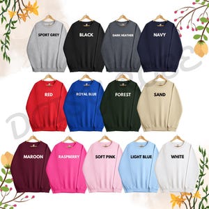 Op de afbeelding: Een selectie van 15 crewneck sweatshirts in verschillende kleuren, waaronder sport grijs, zwart, donker grijs, marineblauw, rood, koninklijk blauw, bosgroen, zand, bordeauxrood, framboos, zacht roze, lichtblauw en wit. Elk sweatshirt heeft een andere kleur en heeft een wit tekstlabel met de kleurnaam.