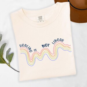 May include: A white t-shirt with the text "Healing is not linear" in navy blue. The text is above a rainbow-colored wavy line.
