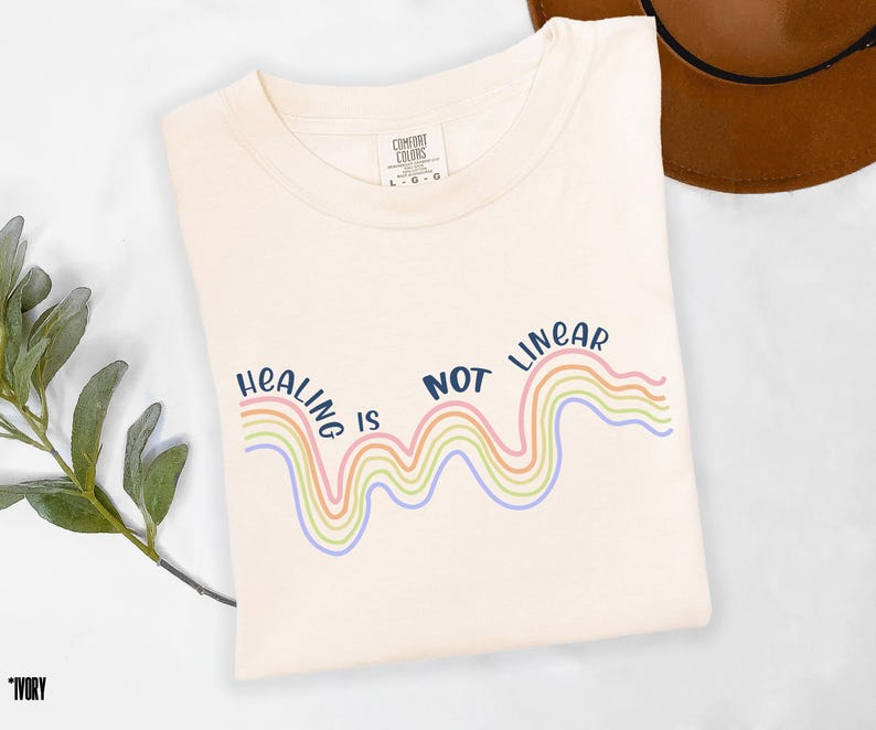 May include: A white t-shirt with the text "Healing is not linear" in navy blue. The text is above a rainbow-colored wavy line.