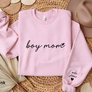 Op de afbeelding: Zachtroze trui met de tekst "boy mom" en een klein hartje. De linkermouw heeft de tekst "mike james" en een hartje.