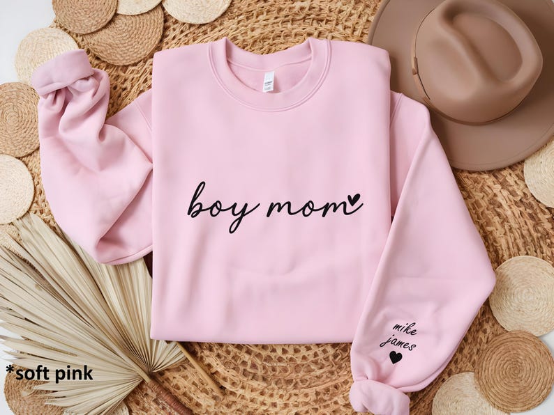 Op de afbeelding: Zachtroze trui met de tekst "boy mom" en een klein hartje. De linkermouw heeft de tekst "mike james" en een hartje.