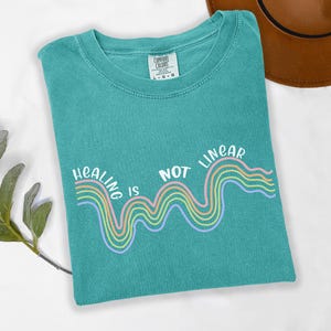 May include: Teal blue t-shirt with a white graphic of a rainbow wave and the text "Healing is not linear".
