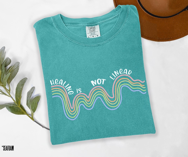 May include: Teal blue t-shirt with a white graphic of a rainbow wave and the text "Healing is not linear".