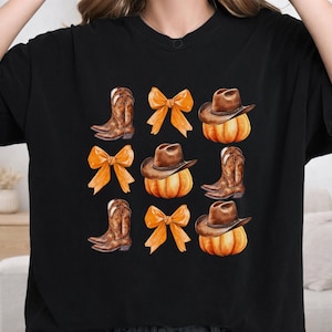 May include: A black t-shirt featuring a repeating graphic print arranged in a grid. The design includes brown cowboy boots, orange bows, and brown cowboy hats resting on top of orange pumpkins, all rendered in a watercolor illustration style.