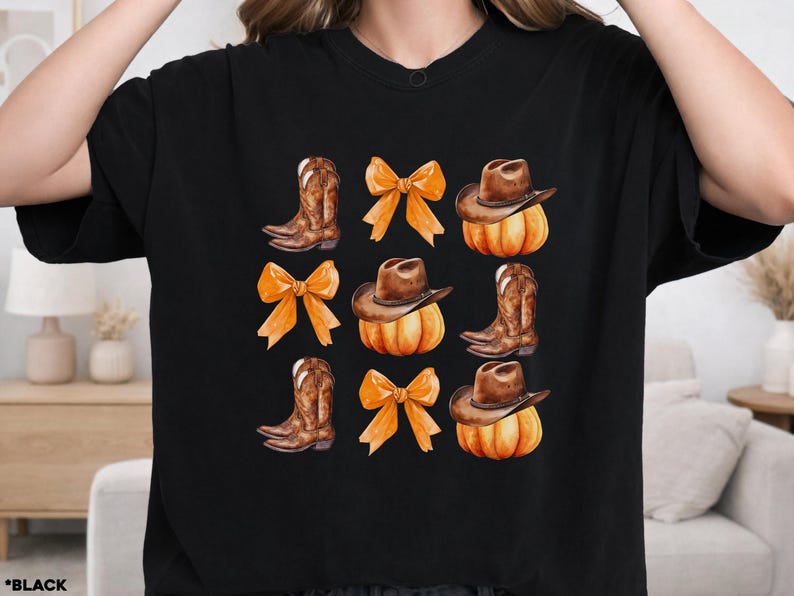 May include: A black t-shirt featuring a repeating graphic print arranged in a grid. The design includes brown cowboy boots, orange bows, and brown cowboy hats resting on top of orange pumpkins, all rendered in a watercolor illustration style.