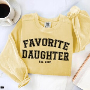 Puede incluir: Una sudadera amarilla mantequilla con las palabras "FAVORITE DAUGHTER" en letras negras, con "EST. 2005" debajo. La camisa de manga larga est&aacute; sobre una superficie blanca con una taza de caf&eacute; y un cuaderno.