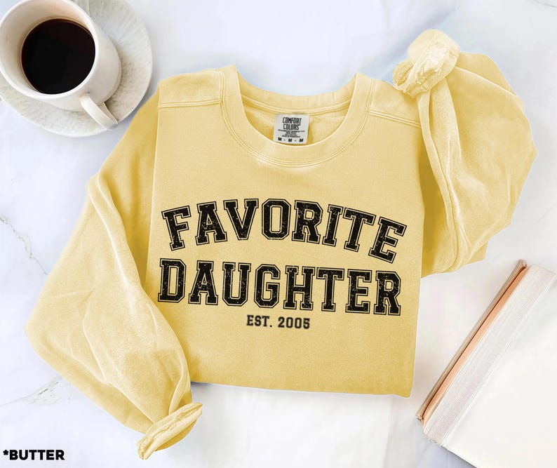 Puede incluir: Una sudadera amarilla mantequilla con las palabras "FAVORITE DAUGHTER" en letras negras, con "EST. 2005" debajo. La camisa de manga larga est&aacute; sobre una superficie blanca con una taza de caf&eacute; y un cuaderno.