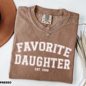 Puede incluir: Camiseta color espresso con las palabras "FAVORITE DAUGHTER" en letras blancas, con "EST. 1996" debajo. Un collar plateado con un peque&ntilde;o colgante descansa sobre la camiseta. Un sombrero marr&oacute;n y una planta seca est&aacute;n en el fondo.