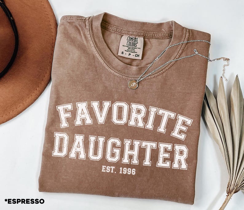 Puede incluir: Camiseta color espresso con las palabras "FAVORITE DAUGHTER" en letras blancas, con "EST. 1996" debajo. Un collar plateado con un peque&ntilde;o colgante descansa sobre la camiseta. Un sombrero marr&oacute;n y una planta seca est&aacute;n en el fondo.