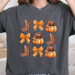 May include: A charcoal gray short-sleeve t-shirt featuring a repeating grid graphic print. The design includes illustrations of brown cowboy boots, orange bows, and brown cowboy hats resting on top of orange pumpkins arranged in three rows.