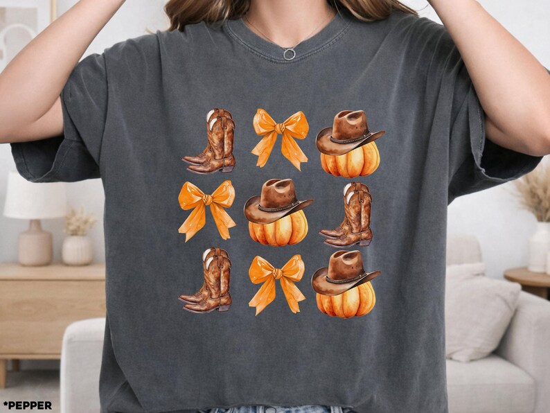 May include: A charcoal gray short-sleeve t-shirt featuring a repeating grid graphic print. The design includes illustrations of brown cowboy boots, orange bows, and brown cowboy hats resting on top of orange pumpkins arranged in three rows.