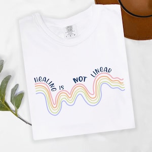 May include: White t-shirt with the text "Healing is not linear" in blue lettering. The text is accompanied by a rainbow-colored wavy line.