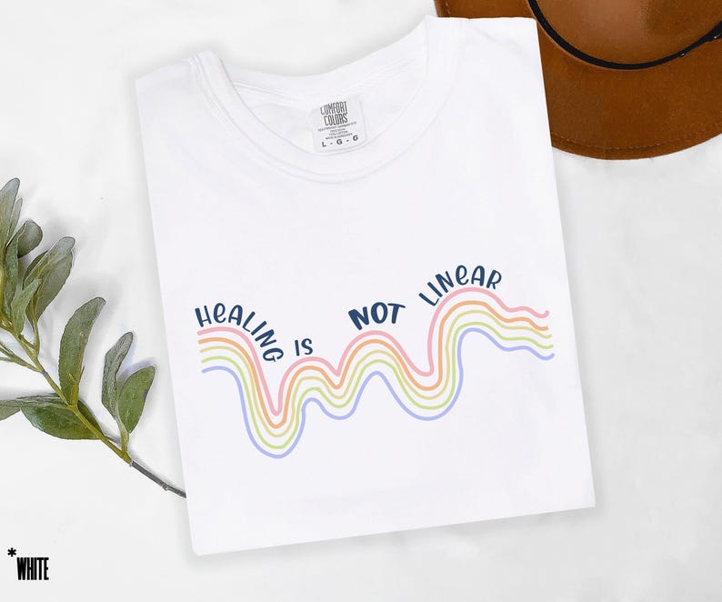 May include: White t-shirt with the text "Healing is not linear" in blue lettering. The text is accompanied by a rainbow-colored wavy line.