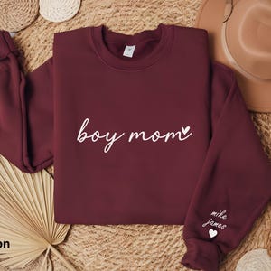 Op de afbeelding: Maroon sweatshirt met de tekst "boy mom" in wit schrift met een klein hartje. De linkermouw heeft de tekst "mike james" in wit met een klein hartje.
