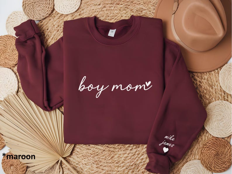 Op de afbeelding: Maroon sweatshirt met de tekst "boy mom" in wit schrift met een klein hartje. De linkermouw heeft de tekst "mike james" in wit met een klein hartje.