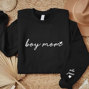 Op de afbeelding: Zwart sweatshirt met de tekst "boy mom" en een klein hartje in witte letters. De rechtermouw heeft de tekst "mike james" en een klein hartje in witte letters.