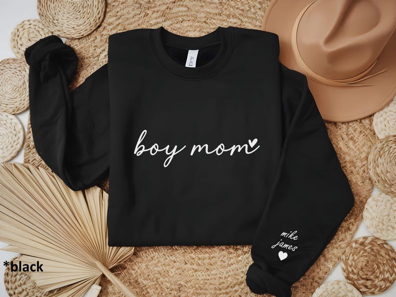 Op de afbeelding: Zwart sweatshirt met de tekst "boy mom" en een klein hartje in witte letters. De rechtermouw heeft de tekst "mike james" en een klein hartje in witte letters.