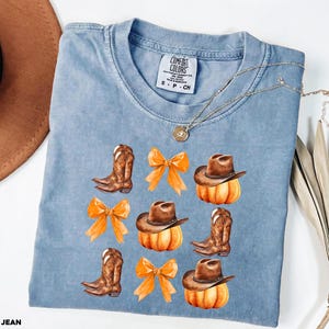 May include: A blue short-sleeve t-shirt with a Comfort Colors label at the neck. The graphic print features brown cowboy boots, orange bows, and brown cowboy hats resting on orange pumpkins arranged in a grid pattern on the front of the shirt.