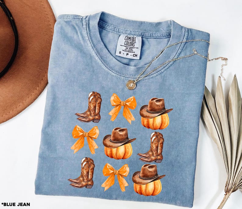 May include: A blue short-sleeve t-shirt with a Comfort Colors label at the neck. The graphic print features brown cowboy boots, orange bows, and brown cowboy hats resting on orange pumpkins arranged in a grid pattern on the front of the shirt.
