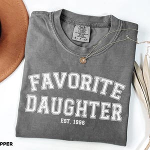 Puede incluir: Una camiseta gris Comfort Colors con las palabras "FAVORITE DAUGHTER EST. 1996" en letras blancas. Un collar plateado con un colgante circular descansa sobre la camiseta doblada. Un sombrero marr&oacute;n y una planta seca en el fondo.