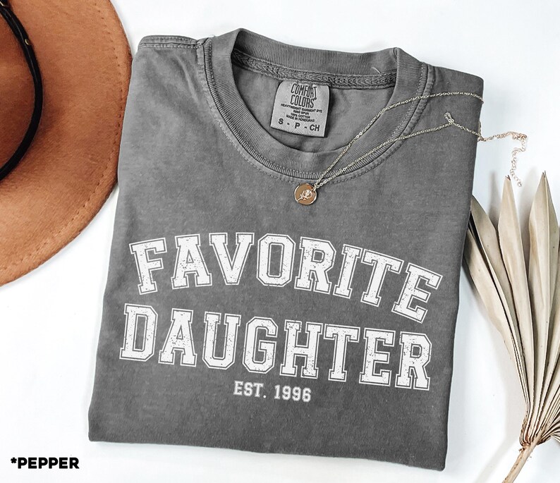 Puede incluir: Una camiseta gris Comfort Colors con las palabras "FAVORITE DAUGHTER EST. 1996" en letras blancas. Un collar plateado con un colgante circular descansa sobre la camiseta doblada. Un sombrero marr&oacute;n y una planta seca en el fondo.