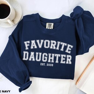 Puede incluir: Una sudadera azul marino con las palabras "FAVORITE DAUGHTER" en letras blancas y en bloque. Debajo est&aacute; "EST. 2005". La sudadera est&aacute; doblada, con una taza de caf&eacute; y un libro en el fondo. Las palabras "TRUE NAVY" est&aacute;n en la parte inferior.