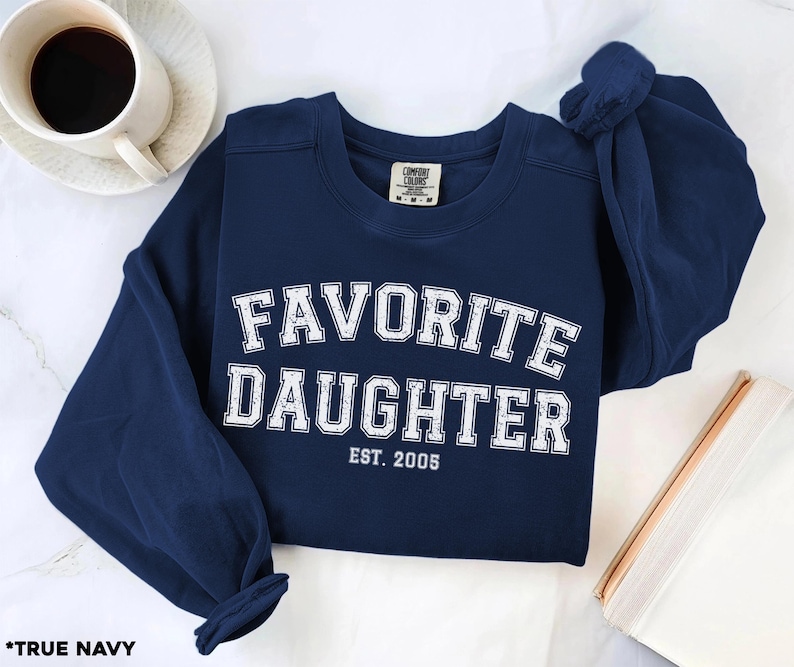 Puede incluir: Una sudadera azul marino con las palabras "FAVORITE DAUGHTER" en letras blancas y en bloque. Debajo est&aacute; "EST. 2005". La sudadera est&aacute; doblada, con una taza de caf&eacute; y un libro en el fondo. Las palabras "TRUE NAVY" est&aacute;n en la parte inferior.