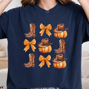 May include: A navy blue short-sleeve t-shirt featuring a repeating grid pattern of brown cowboy boots, orange bows, and brown cowboy hats resting on orange pumpkins. The design is centered on the chest of the shirt, which is labeled as true navy.
