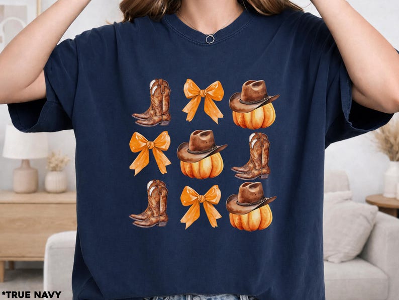 May include: A navy blue short-sleeve t-shirt featuring a repeating grid pattern of brown cowboy boots, orange bows, and brown cowboy hats resting on orange pumpkins. The design is centered on the chest of the shirt, which is labeled as true navy.