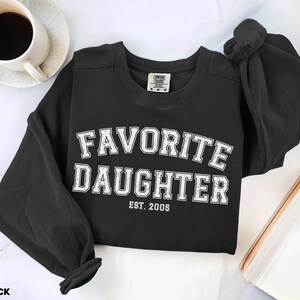 Puede incluir: Sudadera negra con cuello redondo con la frase "FAVORITE DAUGHTER" en letras blancas de estilo universitario. El a&ntilde;o "EST. 2005" est&aacute; impreso debajo. Una taza de caf&eacute; est&aacute; en la esquina superior izquierda. La sudadera est&aacute; doblada.