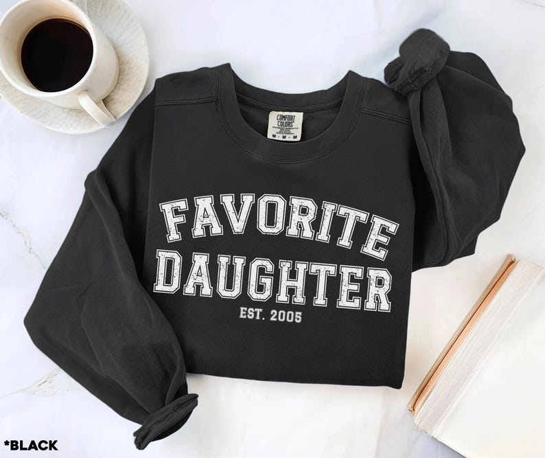 Puede incluir: Sudadera negra con cuello redondo con la frase "FAVORITE DAUGHTER" en letras blancas de estilo universitario. El a&ntilde;o "EST. 2005" est&aacute; impreso debajo. Una taza de caf&eacute; est&aacute; en la esquina superior izquierda. La sudadera est&aacute; doblada.