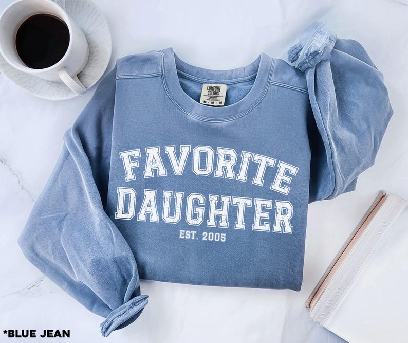 Puede incluir: Una sudadera azul vaquera con las palabras "FAVORITE DAUGHTER" en letras blancas. El texto "EST. 2005" est&aacute; debajo. La sudadera est&aacute; doblada, con una taza de caf&eacute; y un libro en el fondo.