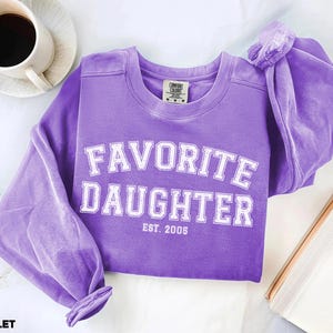 Puede incluir: Una sudadera violeta con las palabras "FAVORITE DAUGHTER" en letras blancas. El texto "EST. 2005" est&aacute; debajo del texto principal. La sudadera es de material suave y tiene mangas largas.