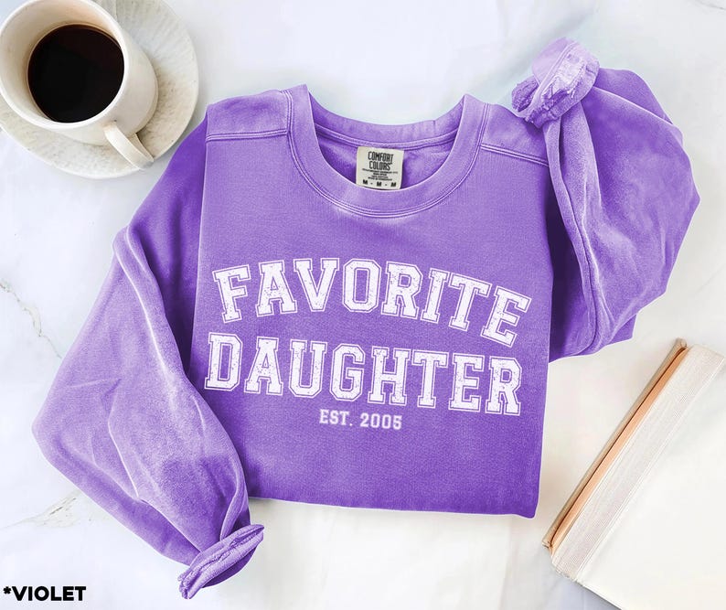 Puede incluir: Una sudadera violeta con las palabras "FAVORITE DAUGHTER" en letras blancas. El texto "EST. 2005" est&aacute; debajo del texto principal. La sudadera es de material suave y tiene mangas largas.