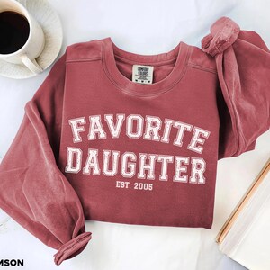 Puede incluir: Sudadera de color carmes&iacute; con las palabras "FAVORITE DAUGHTER" en letras blancas, con "EST. 2005" debajo. La sudadera est&aacute; sobre una superficie blanca con una taza de caf&eacute; y un libro.