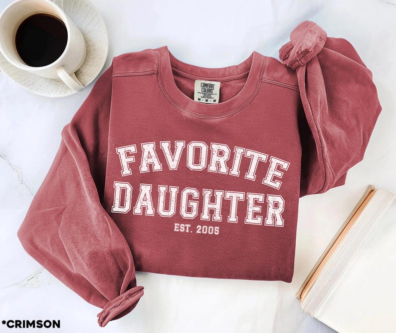 Puede incluir: Sudadera de color carmes&iacute; con las palabras "FAVORITE DAUGHTER" en letras blancas, con "EST. 2005" debajo. La sudadera est&aacute; sobre una superficie blanca con una taza de caf&eacute; y un libro.